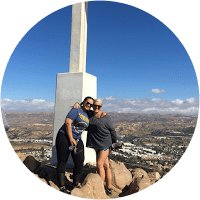 Two women standing on top of a mountain with a cross in the background.